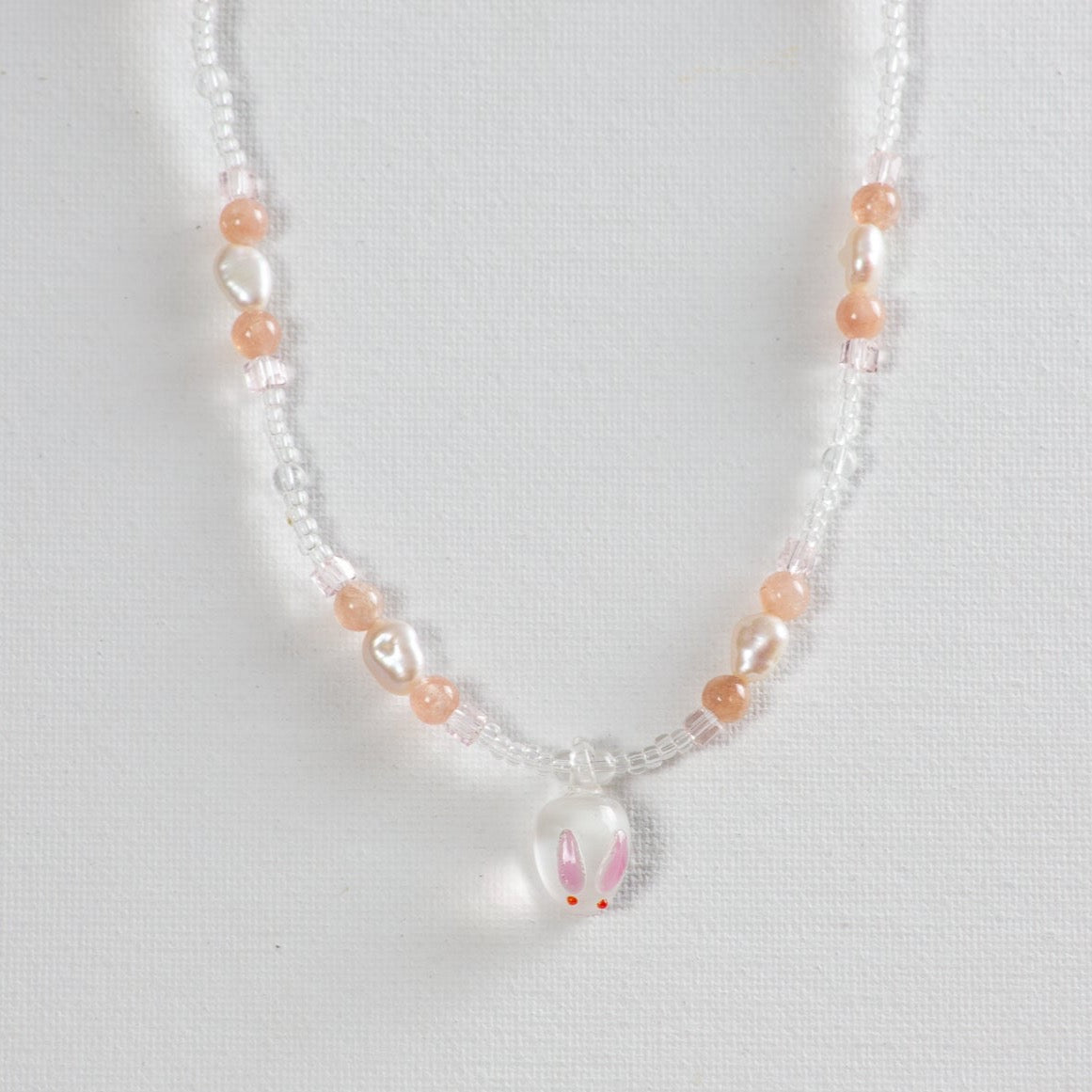 Beaded bunny necklace with clear glass beads forming the main portion, accented by pink glass beads and pearl beads every 2 inches. A charming bunny pendant made of glass hangs at the center. The necklace is placed on a white background.