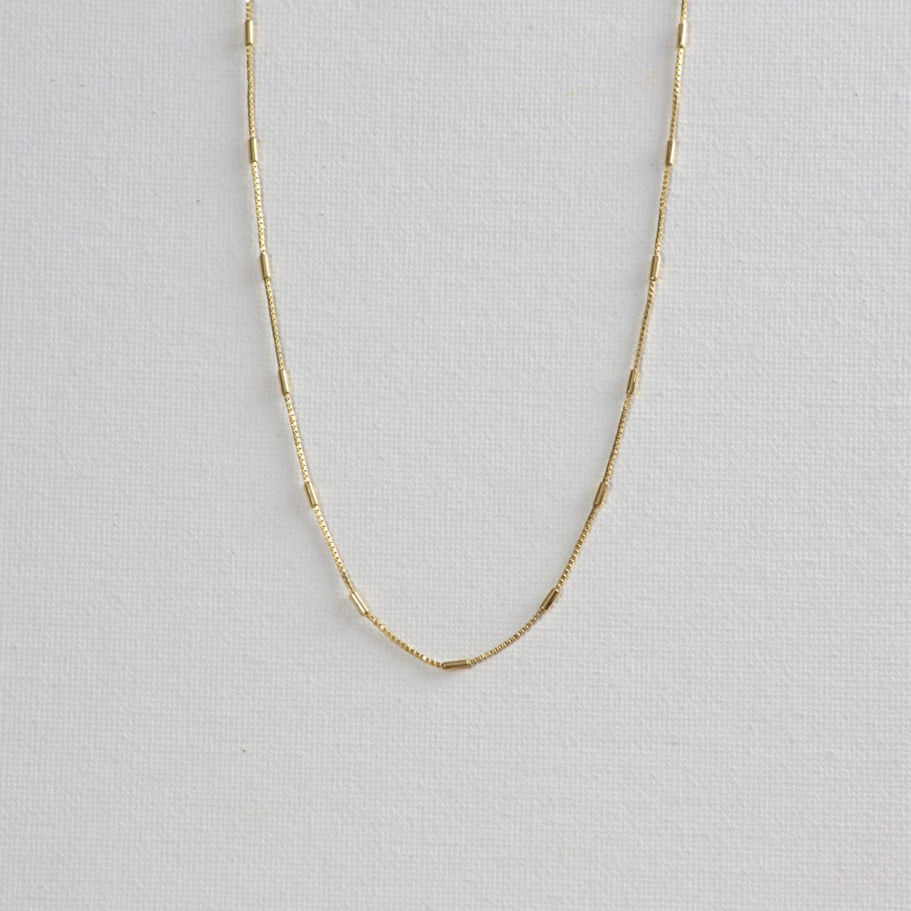 Morse necklace, a gold chain with alternating box chain and tiny gold bar, on a white background.