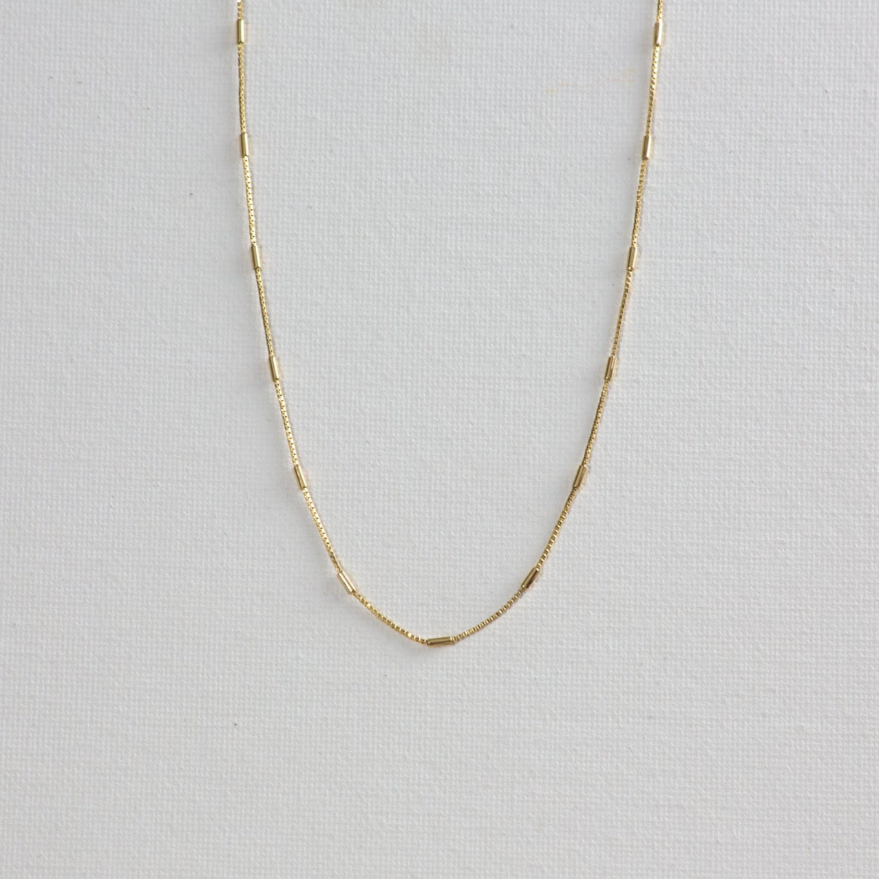 Morse necklace, a gold chain with alternating box chain and tiny gold bar, on a white background.