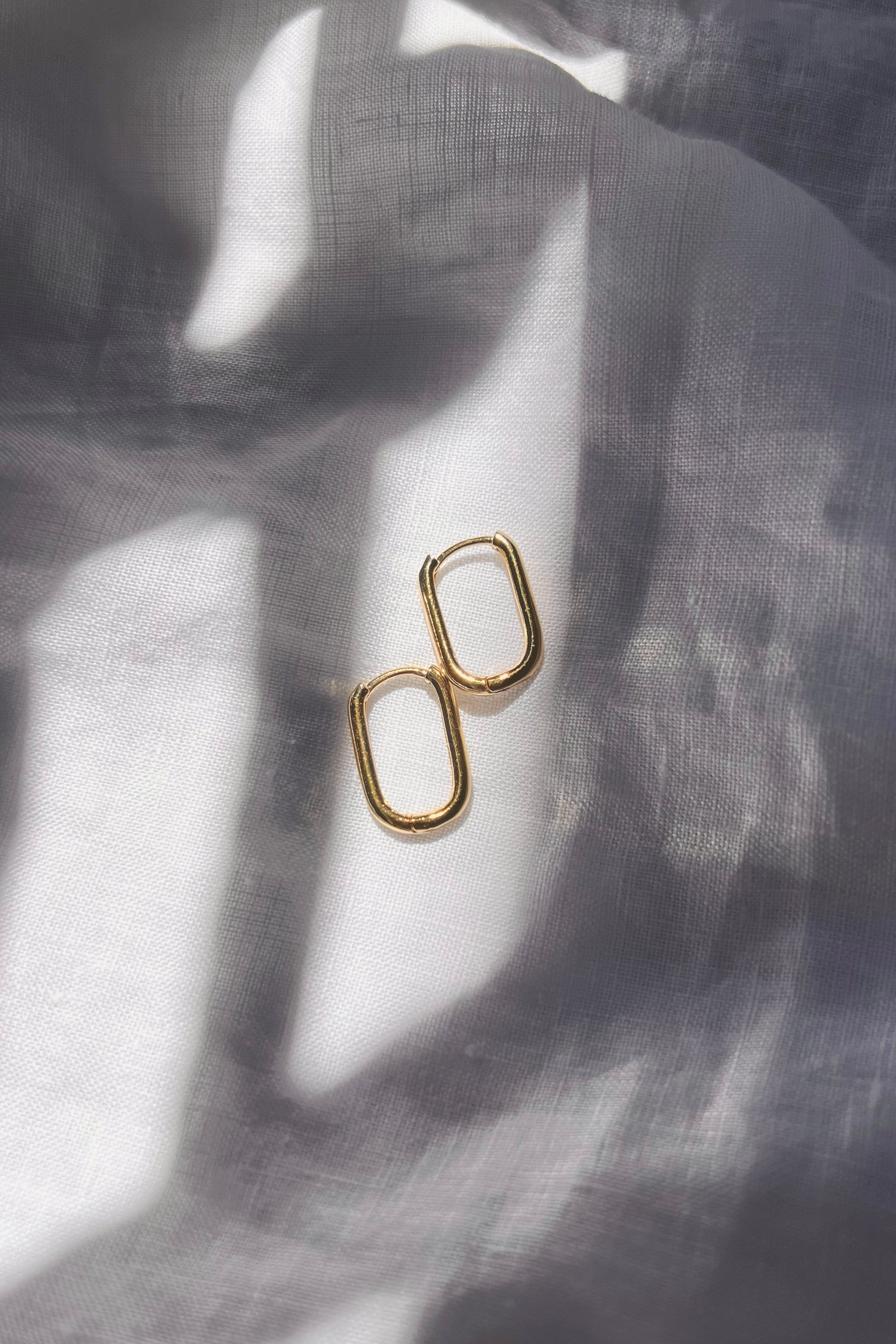 Asa hoops, gold hoops with rounded rectangle shape. The earrings are placed on a white linen fabric with natural sunlight casting over the earrings.