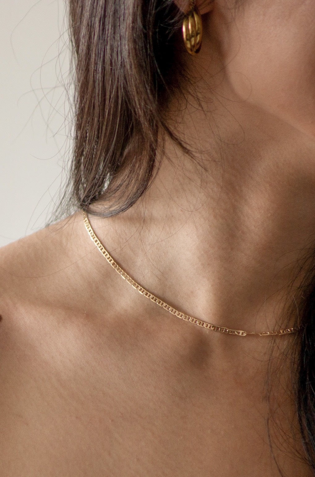 A closer up photo of a model's neck, showcasing the gold chain. In the photo, you can also see the model pairs the necklace with the Enny hoops.